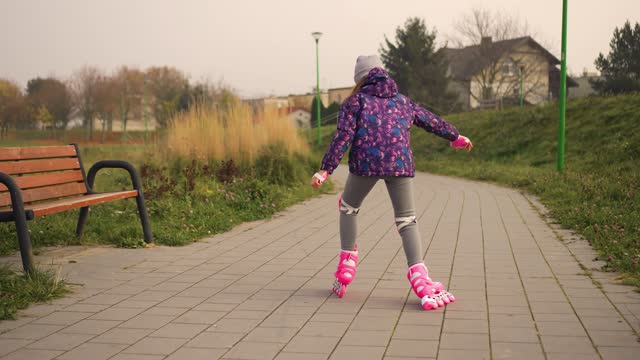 Rear view Little girl riding on roller skate in slow motion outdoors in city park at autumn cloudy day. Kid learning active sport