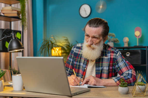 istock Senior businessman working on laptop computer online, writing notes in planner notebook at office 2084051675