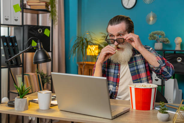 istock Senior man freelancer taking break from work wearing 3D glasses eating popcorn watching movie film 2084051681