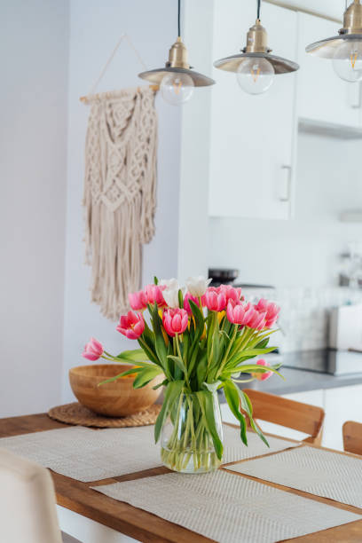 istock Stylish and modern boho, scandi interior of open space white kitchen with tulip flowers in vase on wooden kitchen counter table, macrame, industrial style ceiling lamps. Design home decor. Vertical. 2084062608