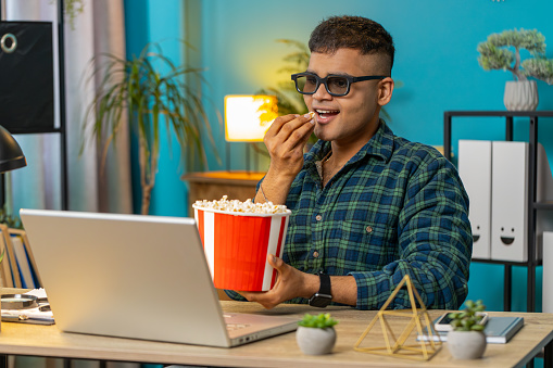 Business Indian man freelancer taking break from work wearing 3D glasses eating popcorn watching movie on laptop. Smiling Arabian guy sitting at home office desk having snacks during break. Lifestyle