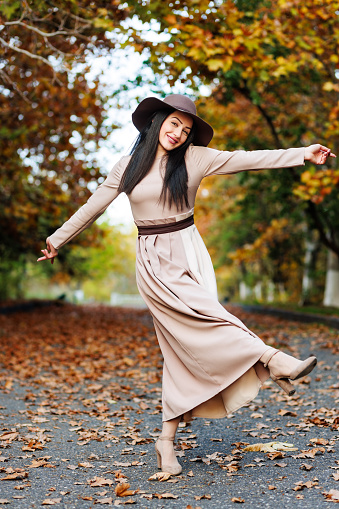 Seasonal joy. A young woman in stylish attire dances along a leaf-covered pathway and celebrating the beauty of autumn