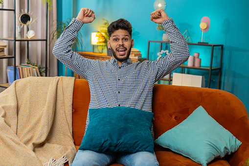 Happy Indian man shouting, celebrating success, winning, goal achievement good news lottery luck victory, watching TV championship at modern home apartment. Arabian guy in living room sits on couch