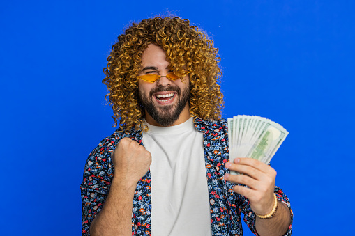 Rich pleased happy man with curly hairstyle wig waving money dollar cash banknotes bills like a fan, success business career, lottery winner, big income wealth. Guy isolated on blue background indoors
