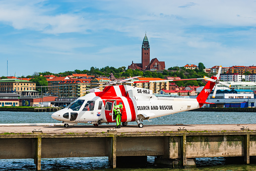 A red and white search and rescue helicopter is parked on top of a pier in Gothenburg, Sweden. The helicopter is ready for action, with its rotors still and its emergency lights flashing.