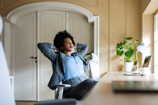 Happy young businesswoman relaxing hands behind head sitting at desk with laptop in office