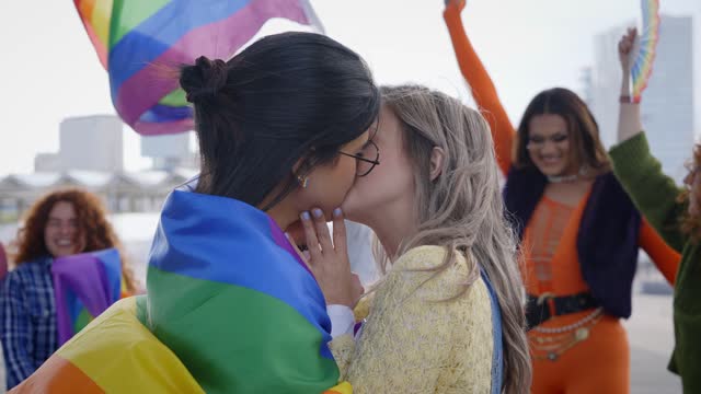 Two young women give each other a tender kiss while carrying a rainbow flag at an LGBT