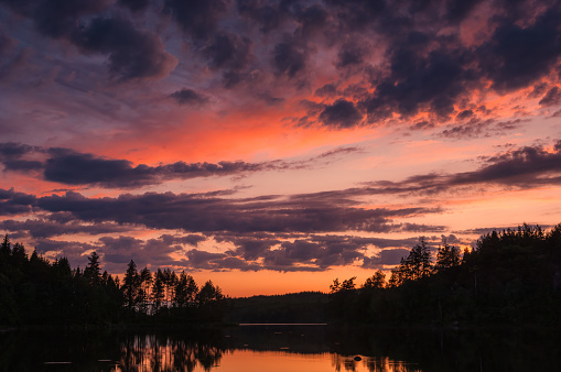 A breathtaking sunset casts a warm glow over a tranquil Swedish lake, with the evening sky painted in shades of pink and orange. Silhouettes of surrounding trees and clouds reflect on the waters surface, creating a serene and picturesque scene.