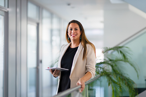 Portrait of a businesswoman looking at a digital tablet while leaning on indoor balcony railing in a modern office with plants.