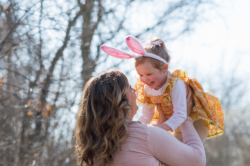 Cute little girl in a yellow dress and a rabbit ears headband is laughing to her mother as she playfully is holding her up outdoors on Easter in the spring.