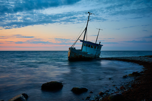 Damaged ship on rocky shore at tranquil beach with idyllic blue cloudy sky in background during sunset