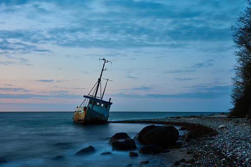 Abandoned ship and rocks on shore at tranquil beach with idyllic cloudy sky in background during sunset