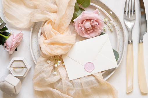 Sealed envelope near cream tulle fabric knot and light pink roses on plates top view, copy space. Wedding stationery mockup with engagement ring in a velvet box. Romantic table place with pastel decor