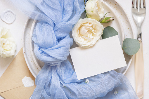 Blank card near light blue tulle fabric and cream roses on plates top view, copy space. Wedding stationery mockup. Romantic table place with horizontal paper card, flowers and pastel decor