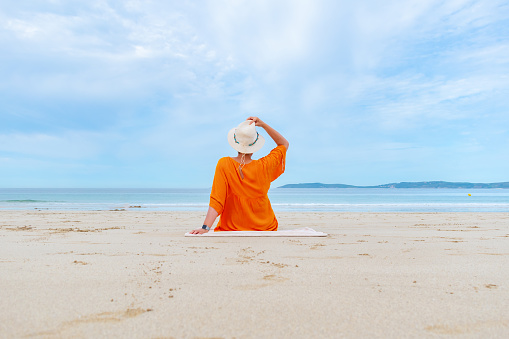 Woman on her back alone on the beach in orange dress and white straw hat looking out to sea.