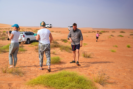 model and photographer on Wahiba desert sand in Oman. They are looking for memory photo with horizon, sand and sky. In foreground, back view of senior woman, while two others, one with camera are walking on sand of  Wahiba desert in Oman. They are looking for memory photo with horizon, sand and sky and parked cars in background.