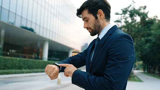 Skilled caucasian business man looking at time while standing at building. Attractive executive manager using smart watch while waiting for partner. Time management, punctuality concept. Exultant.