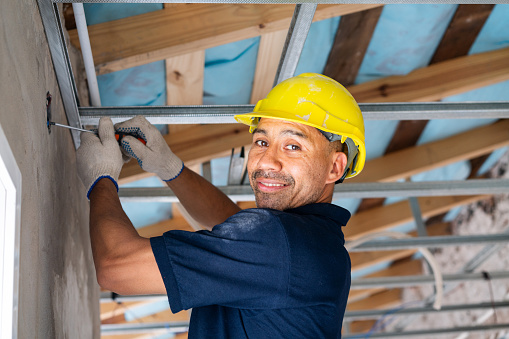 A happy male construction worker in a helmet is focused on installing metal ceiling frames at a building site.