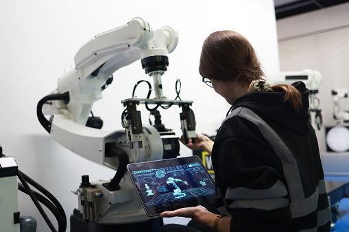 A female mechatronics engineer is working with a robotic arm prototype to examine new programming in the quality room at a manufacturing  welding  factory