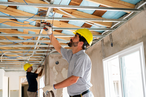 Two construction workers in safety gear are working together to install electrical components in a building's ceiling.