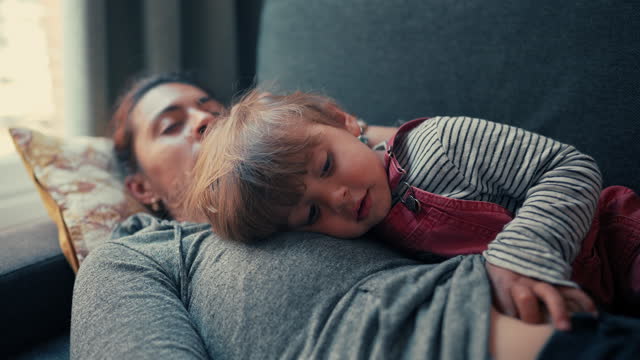 Two year old boy lays on top of mother napping on couch, child feeling mom's heartbeat resting at home, authentic parent asleep and kid gazing with pensive expression. genuine family moment