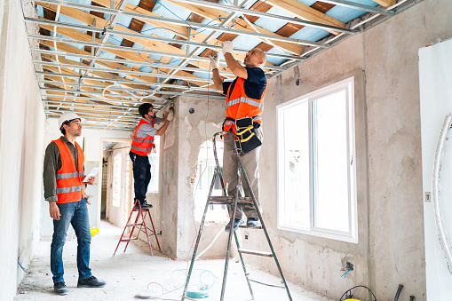 Three construction workers in safety vests and helmets work together installing ceiling framework and windows during an interior renovation project.