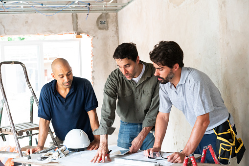 Three construction workers engage in planning and discussion over blueprints on a busy building site.