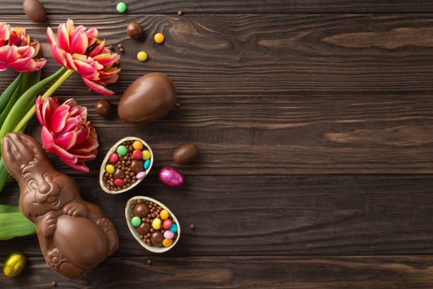 istock Festive Easter scene: Top view of shattered chocolate shells revealing bright sweets, alongside a chocolate hare and fresh tulips on a timber surface, space for copy 2084539337