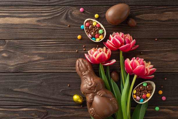 istock Easter treat idea. Top-view photo showcasing smashed chocolate eggs with bright sweets inside, a sweet chocolate hare, and blooming tulips on a wood backdrop, with space for wording or ads 2084539340