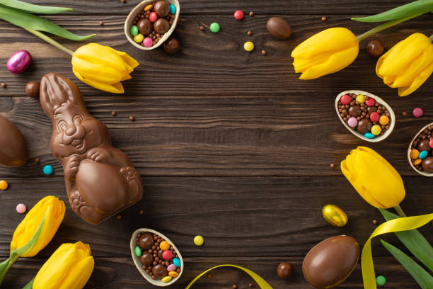 istock Easter gift inspiration. Top view of chocolate eggs cracked open with candies inside, a chocolate bunny, and tulips set on a wooden table, leaving blank space for text or advertisements 2084539346
