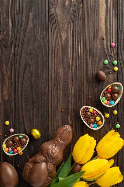 istock Festive Easter candy display: Top view vertical photo of chocolate eggs cracked open, filled with vibrant candies, chocolate rabbit, tulips laid out on wooden table, with space for text or advertising 2084539348
