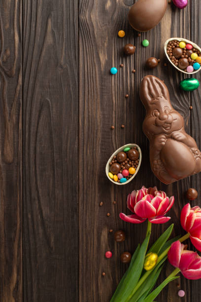 istock Delightful Easter scene featuring overhead vertical shot of broken chocolate eggs overflowing with vibrant candies, chocolate rabbit, and bright tulips on wooden backdrop, leaving space for text or ad 2084539353