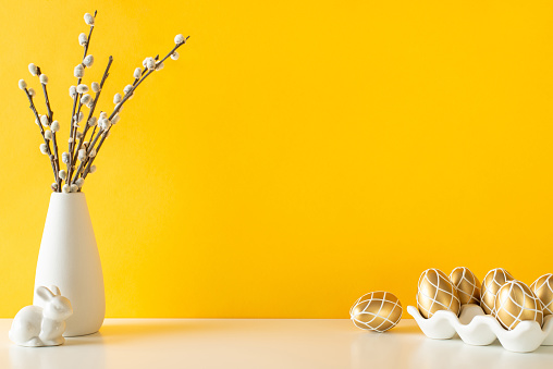 Bright Easter kitchen setup, featuring side view table with stylish ceramic pot for luxurious golden eggs, bunny figure, and pussy-willow in vase, set against lively yellow wall, perfect for messages