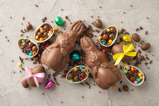 istock Charming Easter set idea: overhead shot of broken chocolate eggs tied with pretty ribbons, brimming with bright sweets, milk chocolate rabbits, candy sprinkles on grey concrete backdrop 2084539371