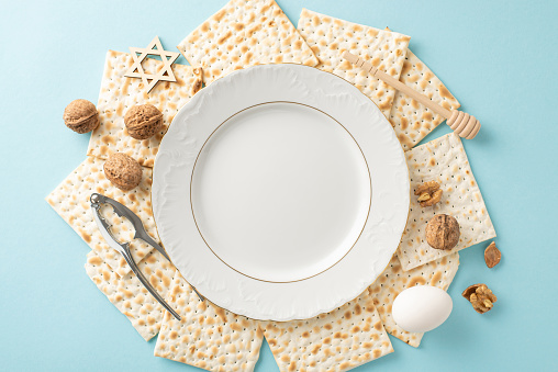 Symbolic Passover arrangement with top view of empty plate, surrounded by matza, walnuts, an egg, a nutcracker, and star of David, laid out on a soft blue background, reserved space for annotations
