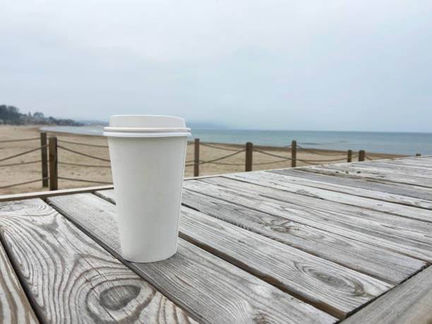 Disposable paper coffee cup on the wooden beach table with foggy beach background. A paper coffee cup with seascape background