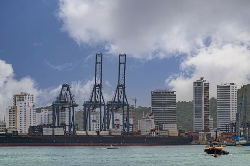 Cartagena, Colombia - July 25, 2023: CMA CGM America container vessel docked at SPRC container terminal under blue cloudscape, tall buildings cityscape in back. Smaller boats on water