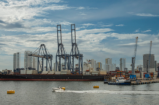 Cartagena, Colombia - July 25, 2023: CMA CGM America docked container vessel at SPRC container terminal under blue cloudscape, tall buildings cityscape in back. Smaller boats on water