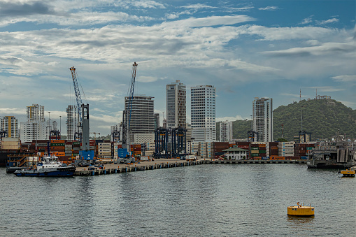 Cartagena, Colombia - July 25, 2023: SPRC container terminal east side pier with 2 cranes in port under blue cloudscape, Smaller boats and stacks of containers. Tall building cityscape