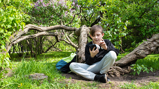 A 10-year-old boy holds a smartphone in his hands, looks at the screen, communicates, in the park.