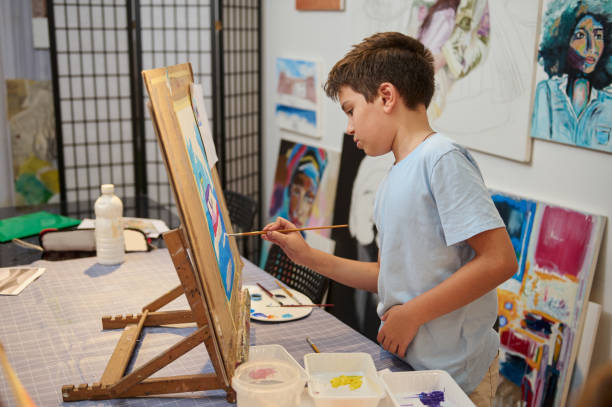 istock Handsome schoolboy drawing picture on canvas, standing by wooden easel in creative workshop. Kids. Education. Art class 2084624881