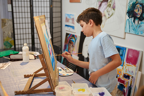 Handsome schoolboy drawing picture on canvas, standing by a wooden easel in creative art workshop. Talented kid, inspired artist painter enjoying his creative hobby indoors, painting with watercolors