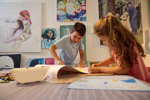 Authentic lifestyle portrait of kids painting and drawing in the visual art class in art school or gallery with displayed artworks on the wall. Kids entertainment and artistic education concept