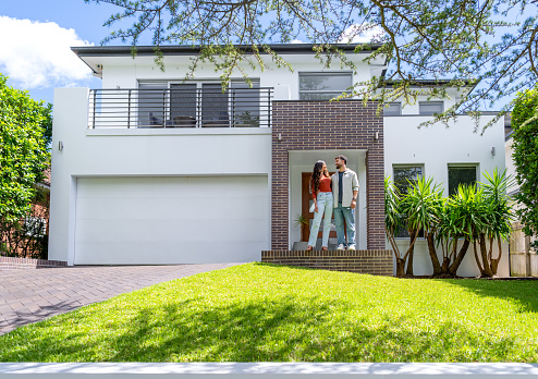 Couple standing proudly in front of their new home. They are both wearing casual clothes. They are looking at each other and smiling. The house is contemporary with a brick facade, driveway, balcony and a green lawn. The front door is also visible. Copy space