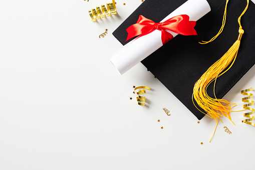 Black mortarboard cap with diploma and gold tinsel on white background. Flat lay graduation party concept.