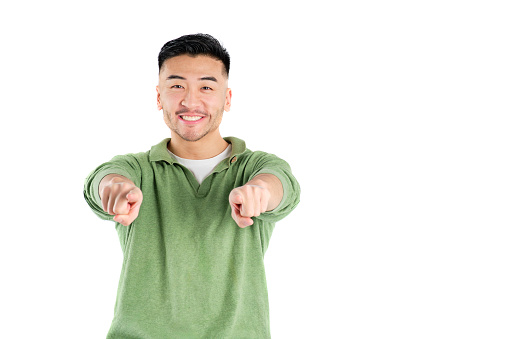 smiling asian young man in green shirt with arms outstretched pointing at camera on white background