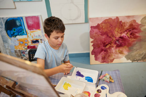 istock Top view Caucasian elementary age school student boy holding paintbrush and mixing watercolors on a palette at art class 2084674541
