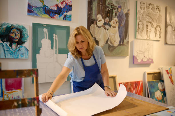 istock Confident female art class teacher rolling out a white design paper sheet on the table, working in her creative workshop 2084692363