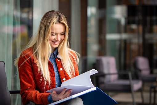 In this close-up capture, a delighted student sits engrossed in her reading, adorned in a vibrant red jacket. With a beaming smile, she exudes happiness amidst her studies. This image, set against modern architecture, portrays the sheer joy of learning. Perfect for educational promotions and uplifting visual content.