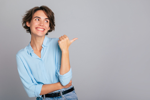 Shot of excited happy female in blue shirt demonstrating and pointing on copy space adverts isolated over gray background